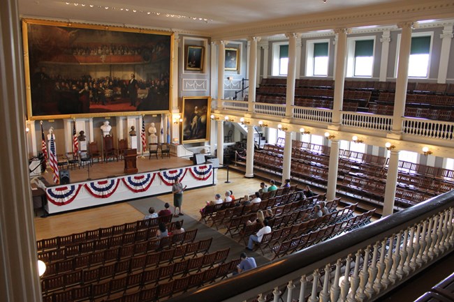 The Great Hall of Faneuil Hall is an open meeting space with a gallery level. Doric and Ionic columns support the room. Portraiture and busts decorate the space.