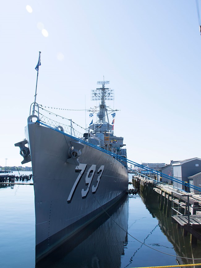 USS CASSIN YOUNG is a steel warship shown here moored alongside a pier. The identification numbers 7 9 3 are painted on the ship's hull. Her jack staff is flying a 50 star jack on the bow. On the tower of the ship are signal flags and the ship's RADAR.