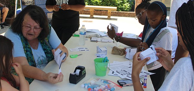 A volunteer shows a group of children how to build a rustic log cabin out of paper.