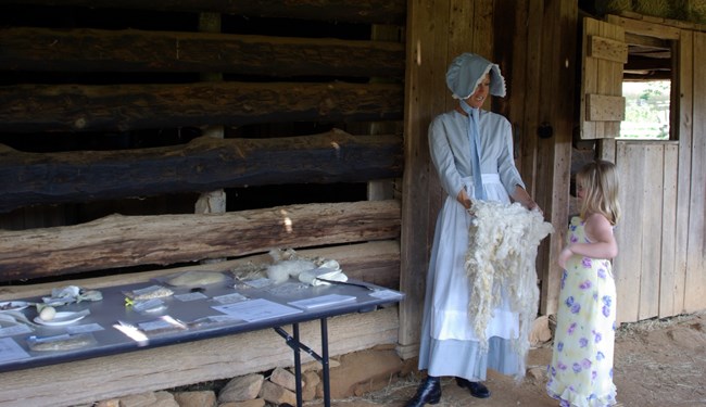 A female in period clothing with a sheepskin stands next to a young female child.