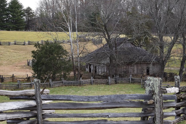 A rustic fence in the foreground with a barn and a horse peeking out of his stall in the background.
