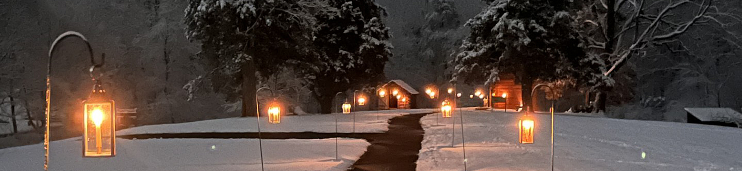 A snow-covered nighttime scene with lighted lanterns leading down a path with a pair of rustic cabins in the distance.