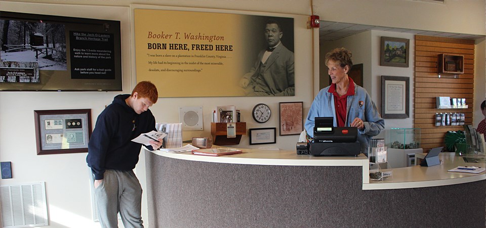A photograph of a visitor reading while standing next to a park service volunteer standing at the cash register.