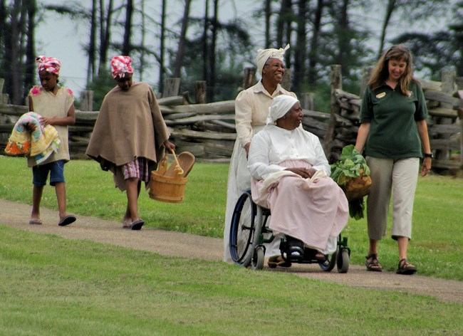 A volunteer and a group of reenactors with a wheelchair walk along a path.