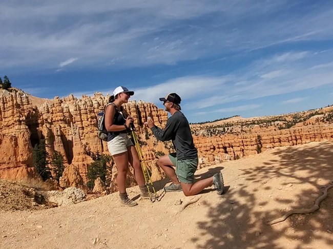 A man kneels as he proposes to a woman along a red rock trail.