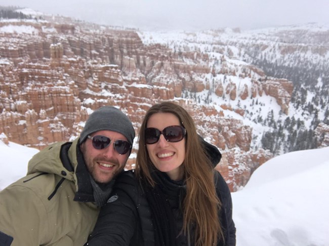 A man and a woman in winter clothing stand at the edge of a snowy cliff.