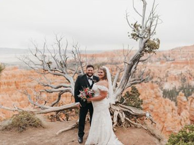 A man in a suit and a woman in a wedding dress hold each other in front of a red rock landscape below them