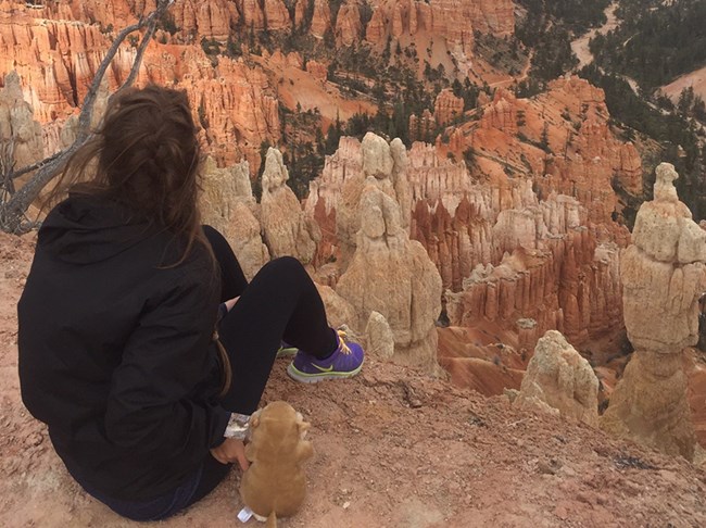 A woman sits with a small stuffed animal at the edge of a red rock cliff.