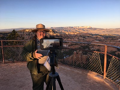 Ranger stands in front of phone on tripod presenting a virtual program at sunset