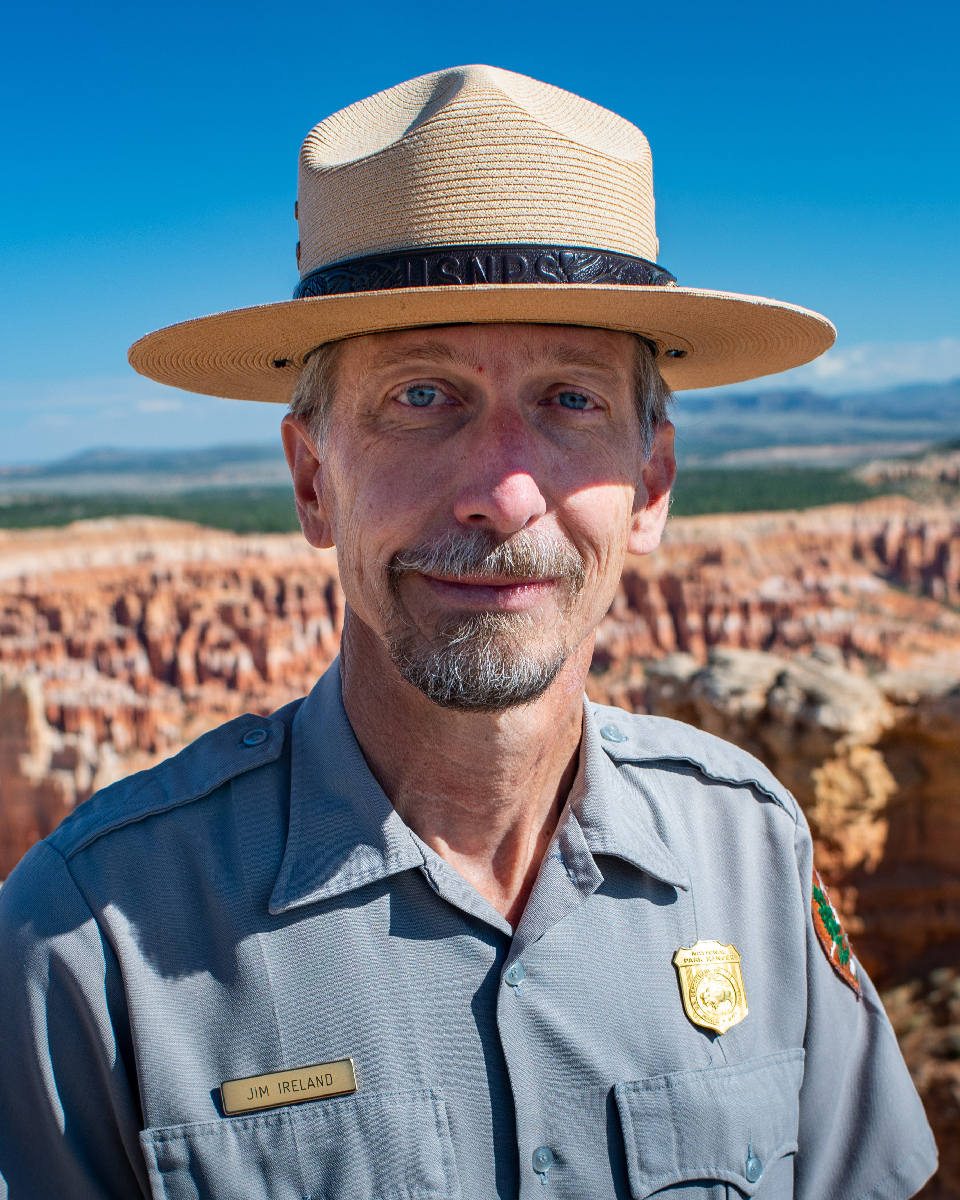 Man with beard and ranger flat hat stands in grey uniform shirt with background of vast redrock landscape
