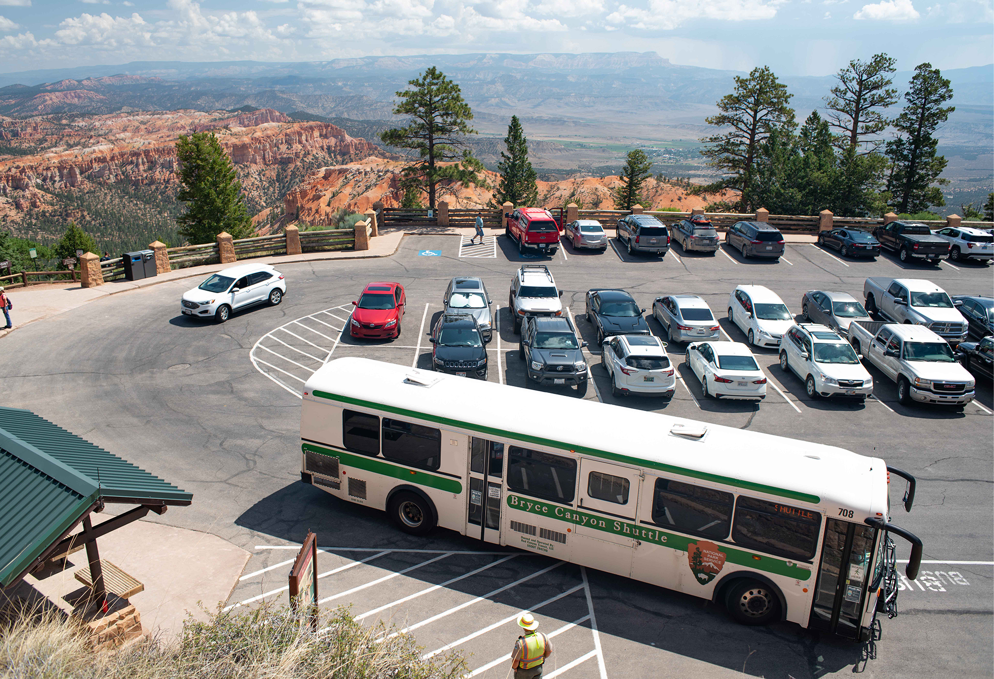 A white and green shuttle bus drives through a busy parking lot in a forested, red rock landscape.