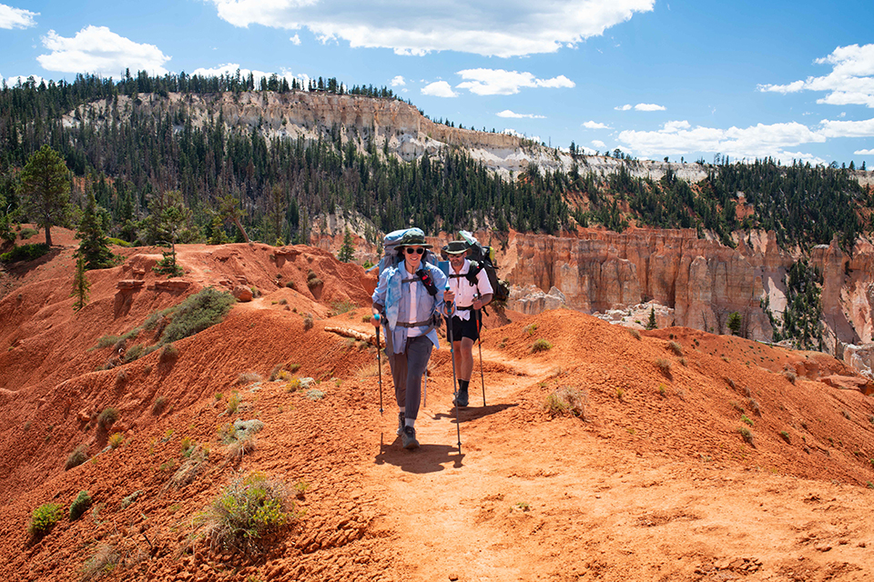Two hikers travel along red rock trail lined with trees and distant cliffs
