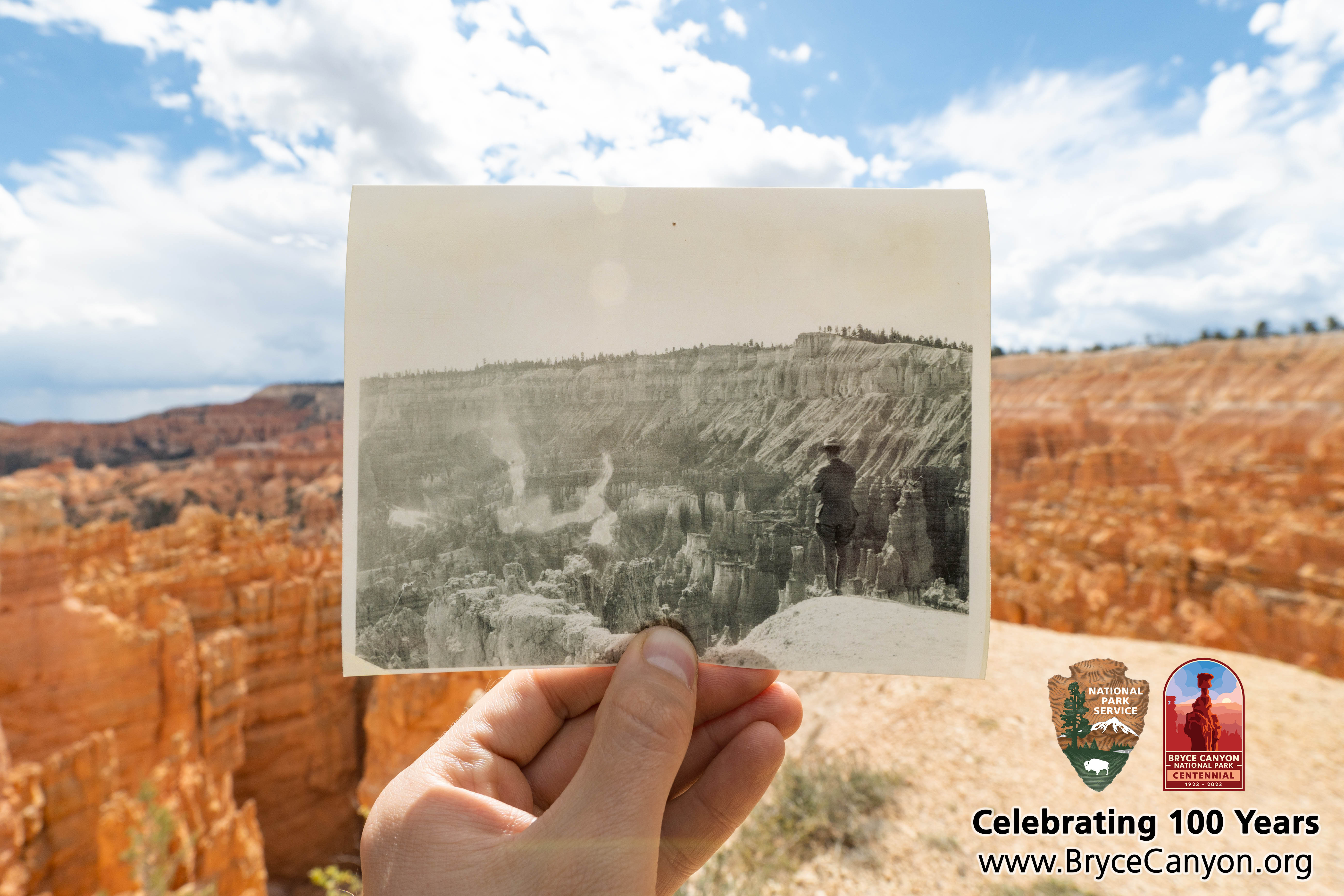 A hand holds up a black and white photo of a man at the edge of a cliff superimposed on a colorful landscape of red rock spires and cliffs.