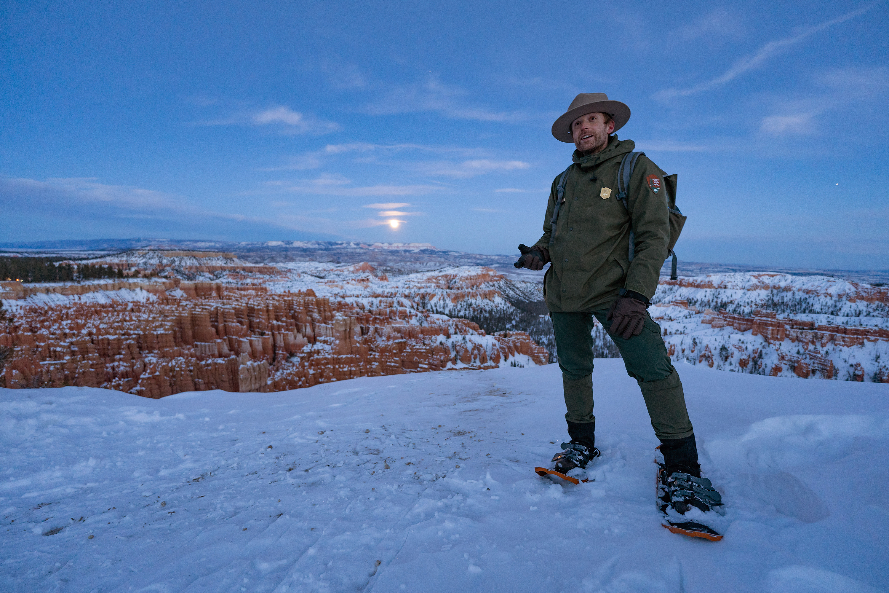 A ranger stands in the snow wearing snow shoes with a bright full moon behind him.