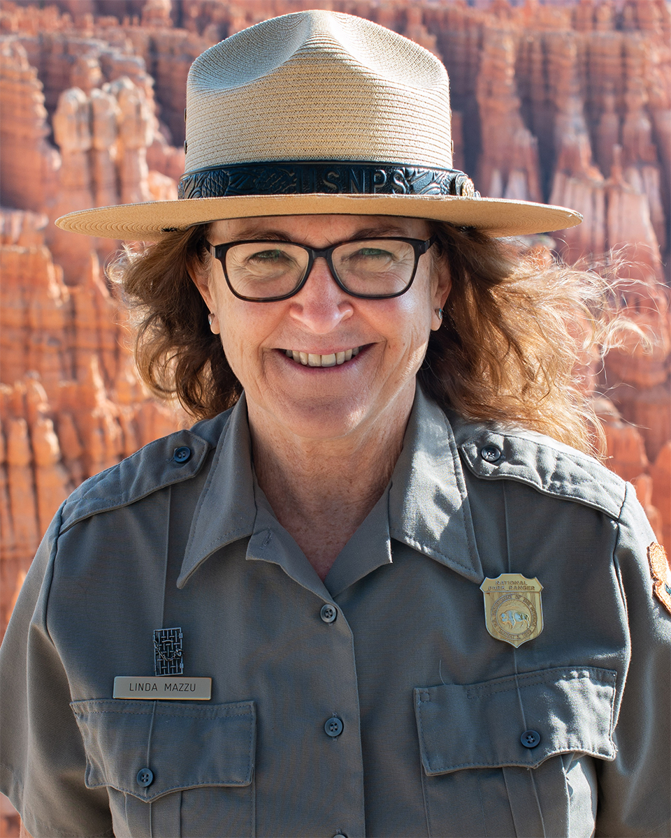 Linda Mazzu Photo Woman wearing glasses and ranger uniform smiles with background of red and white limestone spires