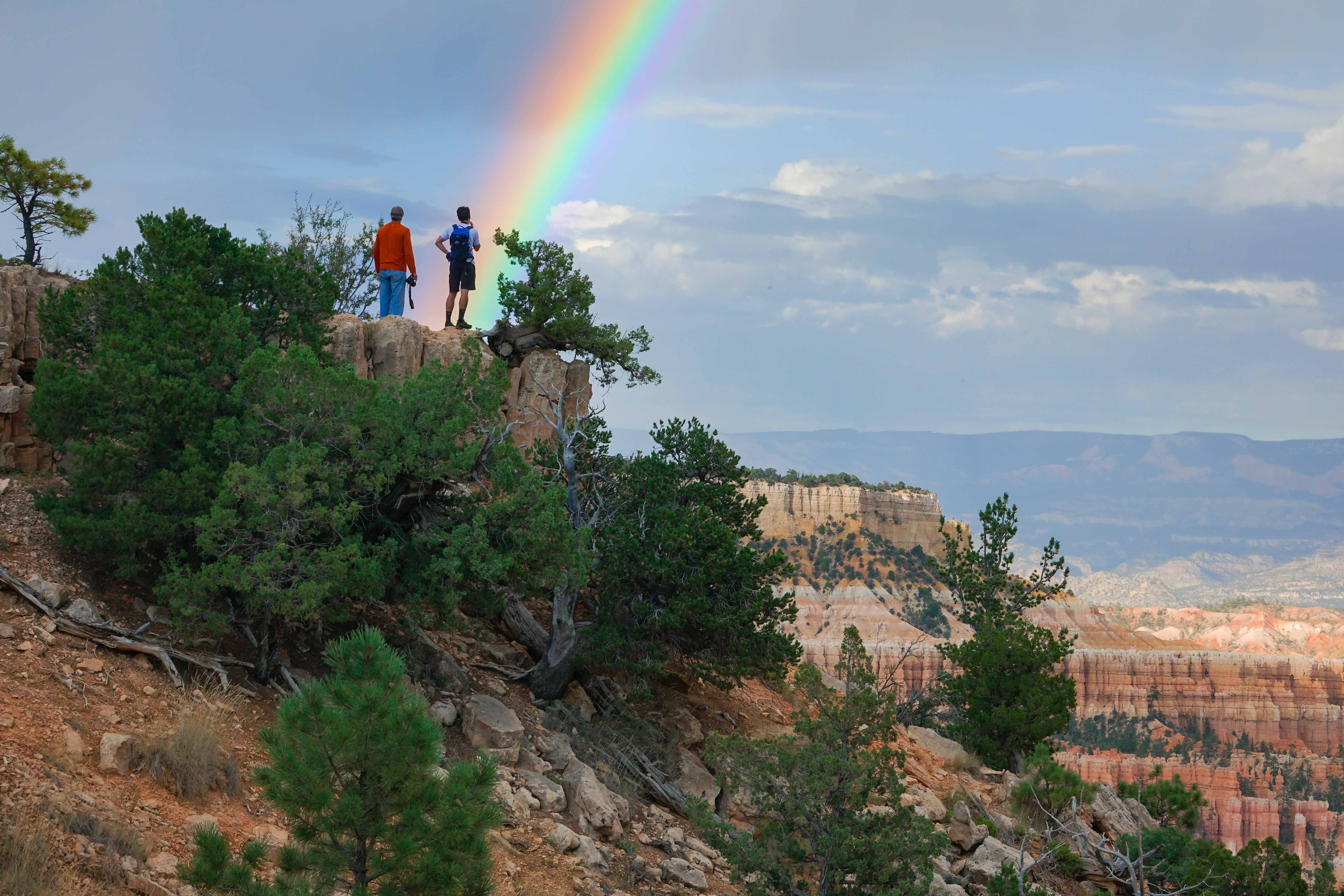 Two people standing along the rim trail with a large rainbow in the sky