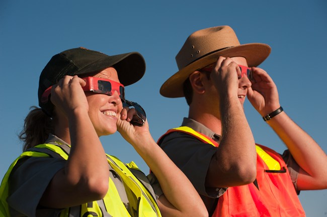 Two rangers in bright yellow and orange vests use special glasses to view the eclipse
