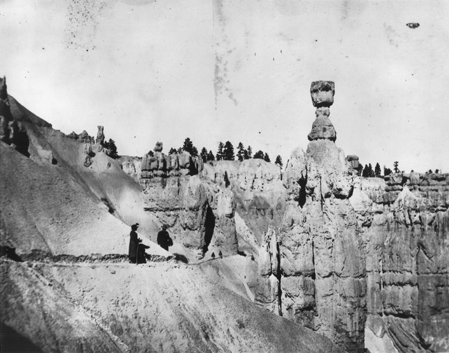 A black and white photo of a large columnular rock formation with a woman in a dress standing on a nearby trail.