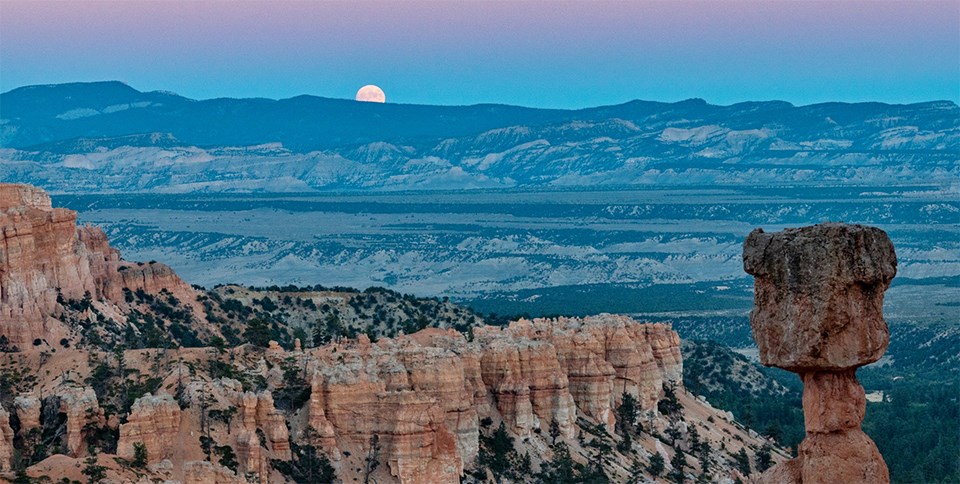 A full moon rises along a distant horizon beyond a rock spire known as Thors Hammer and other red rock features