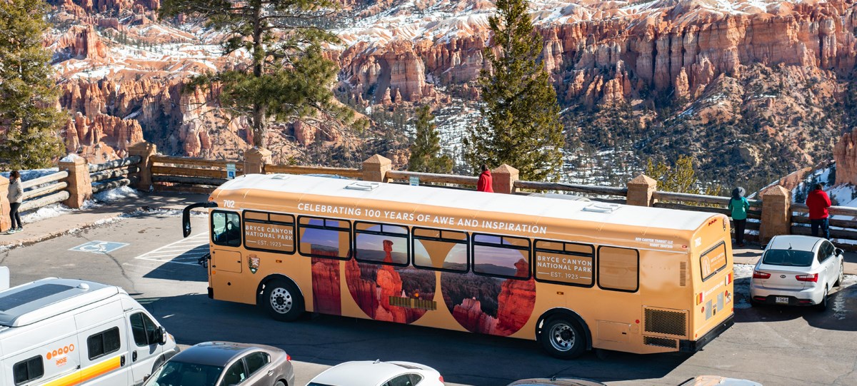 A tan colored shuttle bus can be seen against a vast backdrop of red rocks