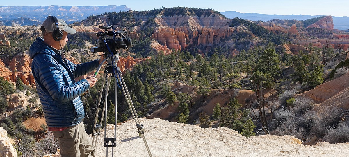 A man with a camera on a tripod views a vast landscape of red rock cliffs, spires, and forest.