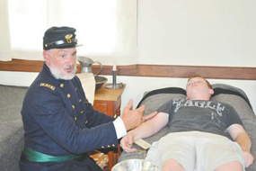 Tim Fields dressed in historic military uniform demonstrates to Ryan Kelly, lying down on a bed, how doctors treated soldiers during the Civil War.