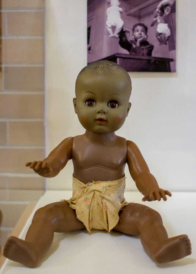 Small brown plastic doll in front of photo of child being presented with white and brown dolls.