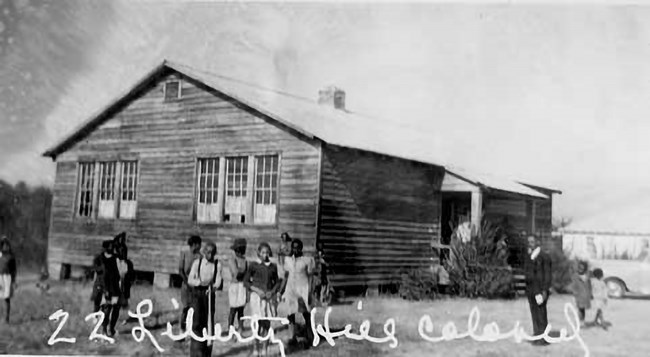 African American students and teacher standing in front of a wood and tar paper shack with text that reads Liberty Hill Colored School.