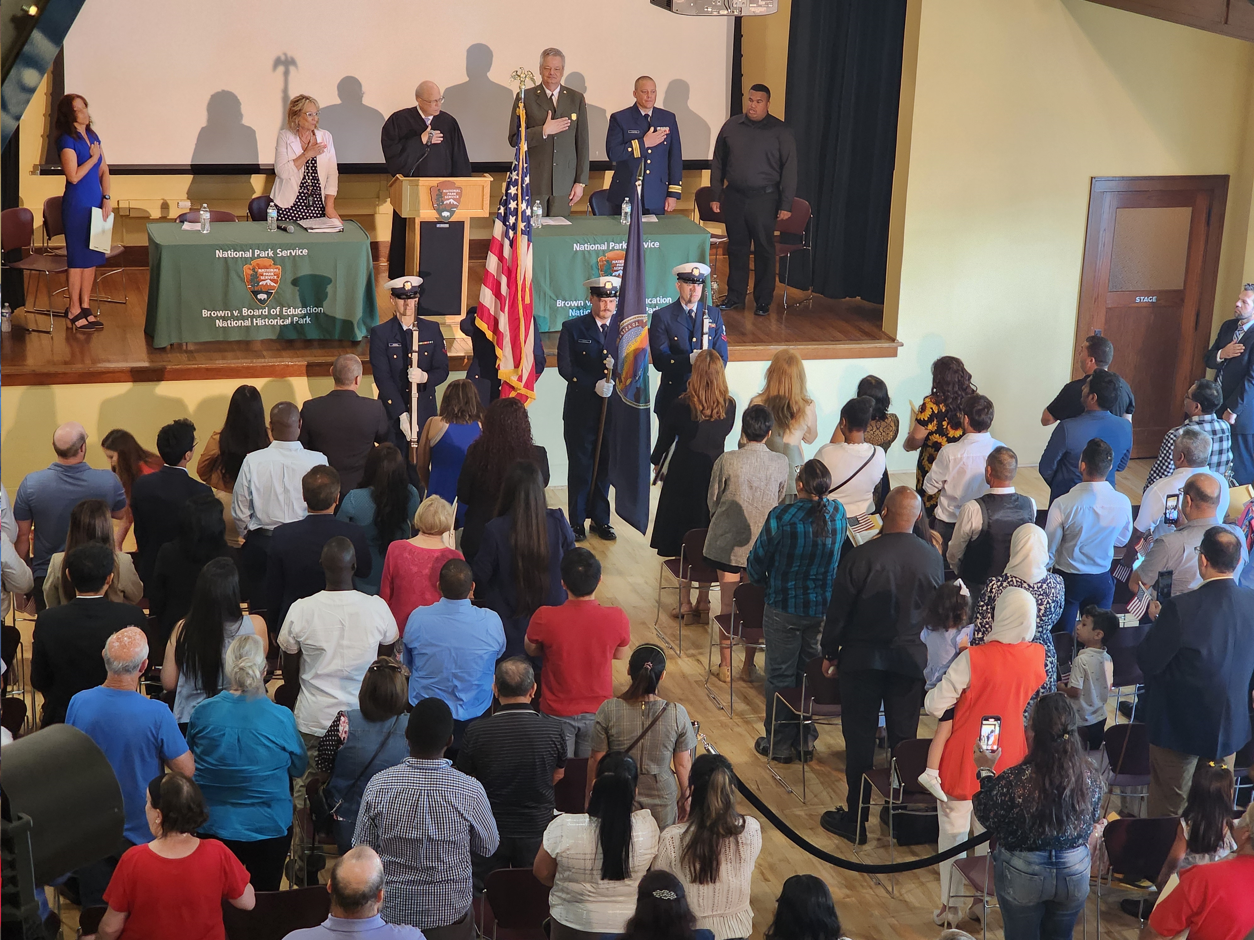 Naturalization Ceremony at Brown v. Board of Education NHP Attendees of a naturalization ceremony stand and place their hands over their hearts in an auditorium with wooden floor.