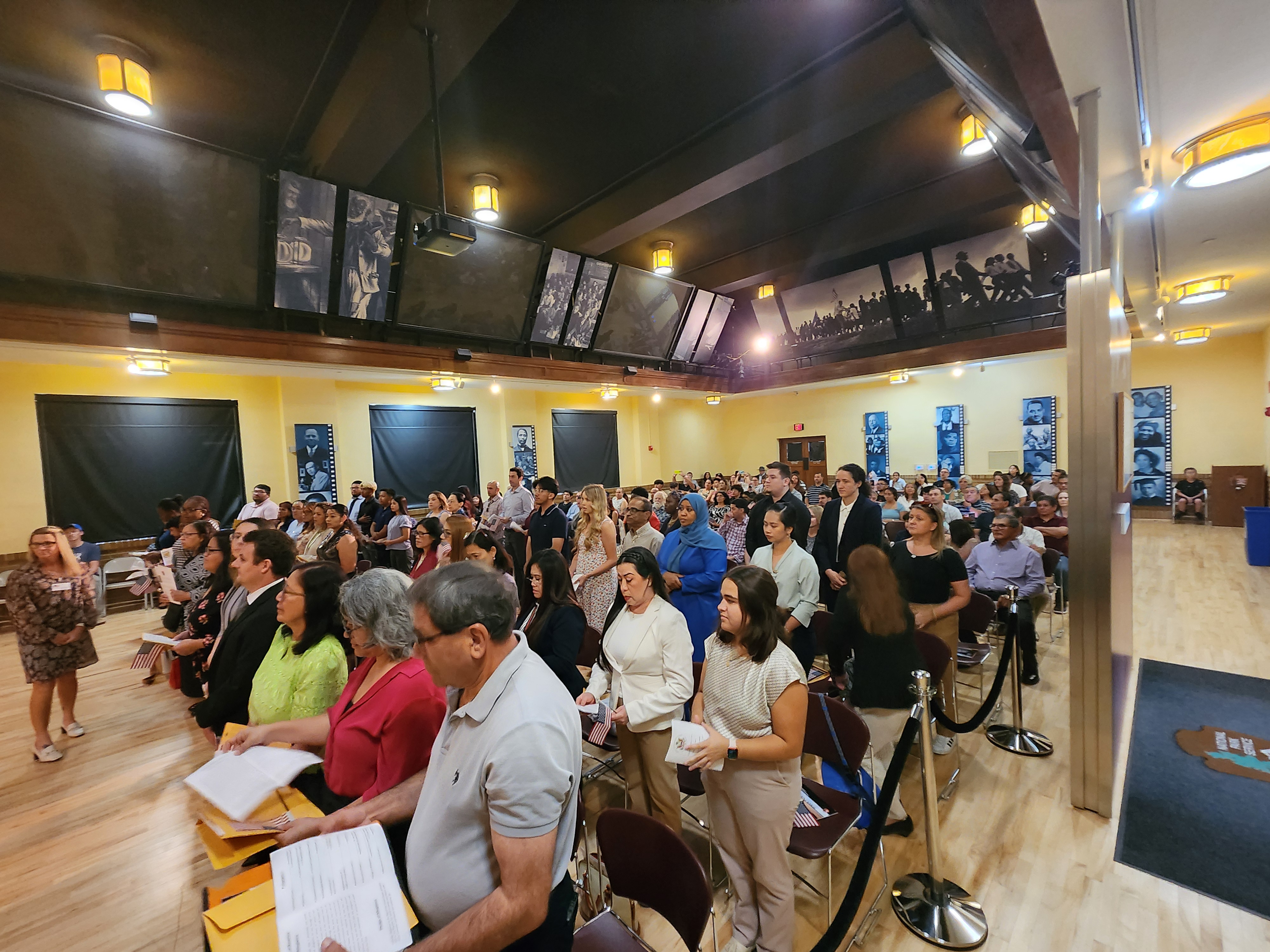 20240725_101127 People stand in an auditorium surrounded by friends and family while holding folders of documents.