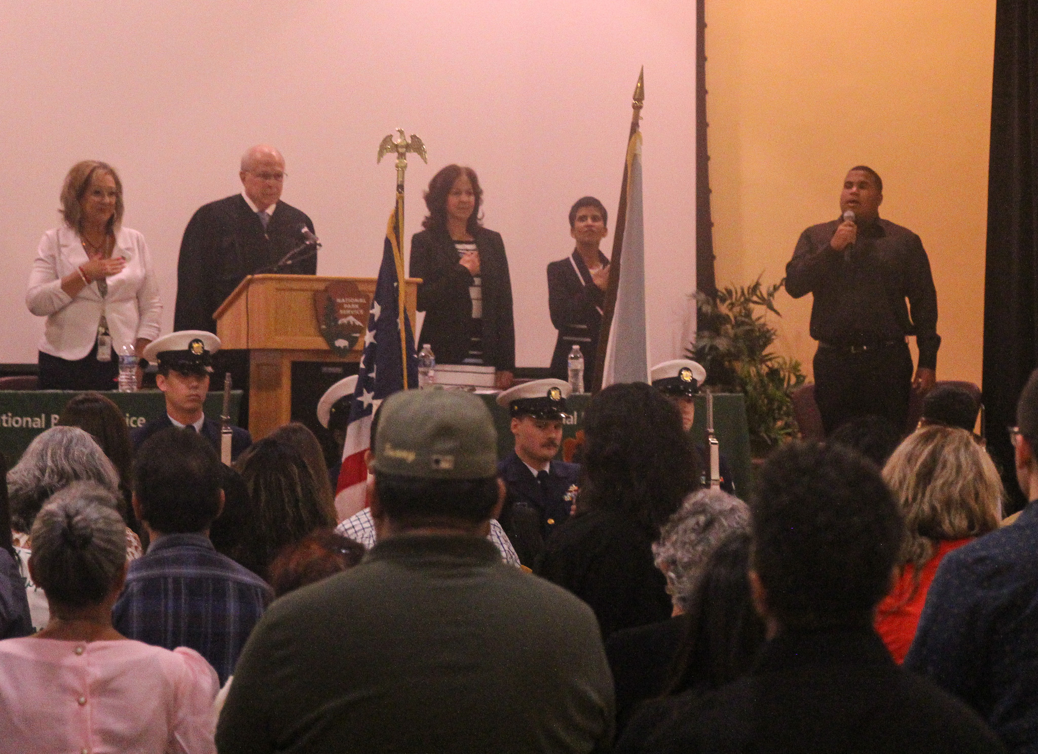 3rd Naturalization Ceremony Five people can be seen standing on the top of a stage with their hands over their heart pledging allegiance to the United States flag. A large crowd can be seen looking towards the stage and pledging allegiance to the United States flag.