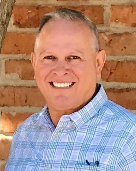DennisVasquez Headshot of a middle aged white man with white and gray hair in front of a brick wall while wearing a blue and gree plaid shirt