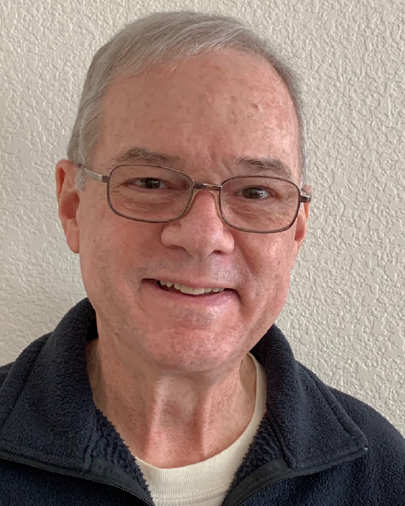 StephenAdams An older white man with white hair and metallic rimmed glasses in a white shirt and blue sweater smiles in front of a blank white wall