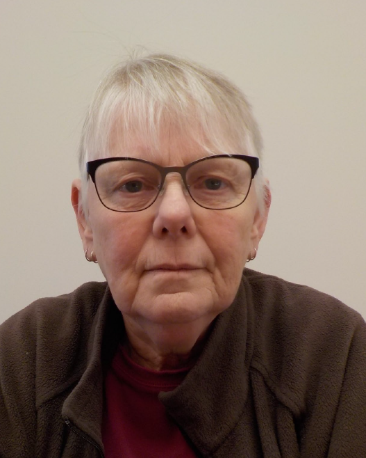 TheoraMcVay Headshot of an older white woman with short white hair, black glasses a red shirt and brown sweater