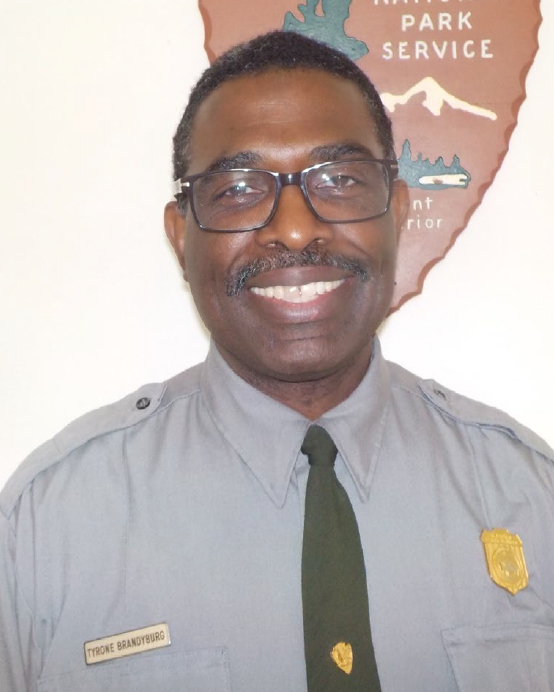 TyroneBrandyburg Headshot of a middle aged Black male park ranger with black and gray short hair, black glasses and a mustach smiling in his green and grey service uniform