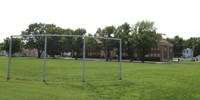 Photo of historic baseball backstop in the foreground of a photo depicting the brick Monroe elementary school building in the summer with a green grass field and trees blocking much of the building.