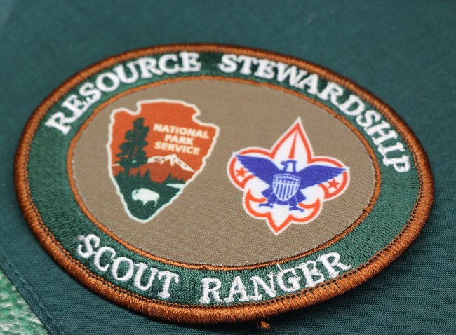 A oval patch with a green ring along the edge which says "Resource Stewardship Scout Ranger" The ring surrounds a smaller brown oval that has the NPS arrowhead and boy scout logo.