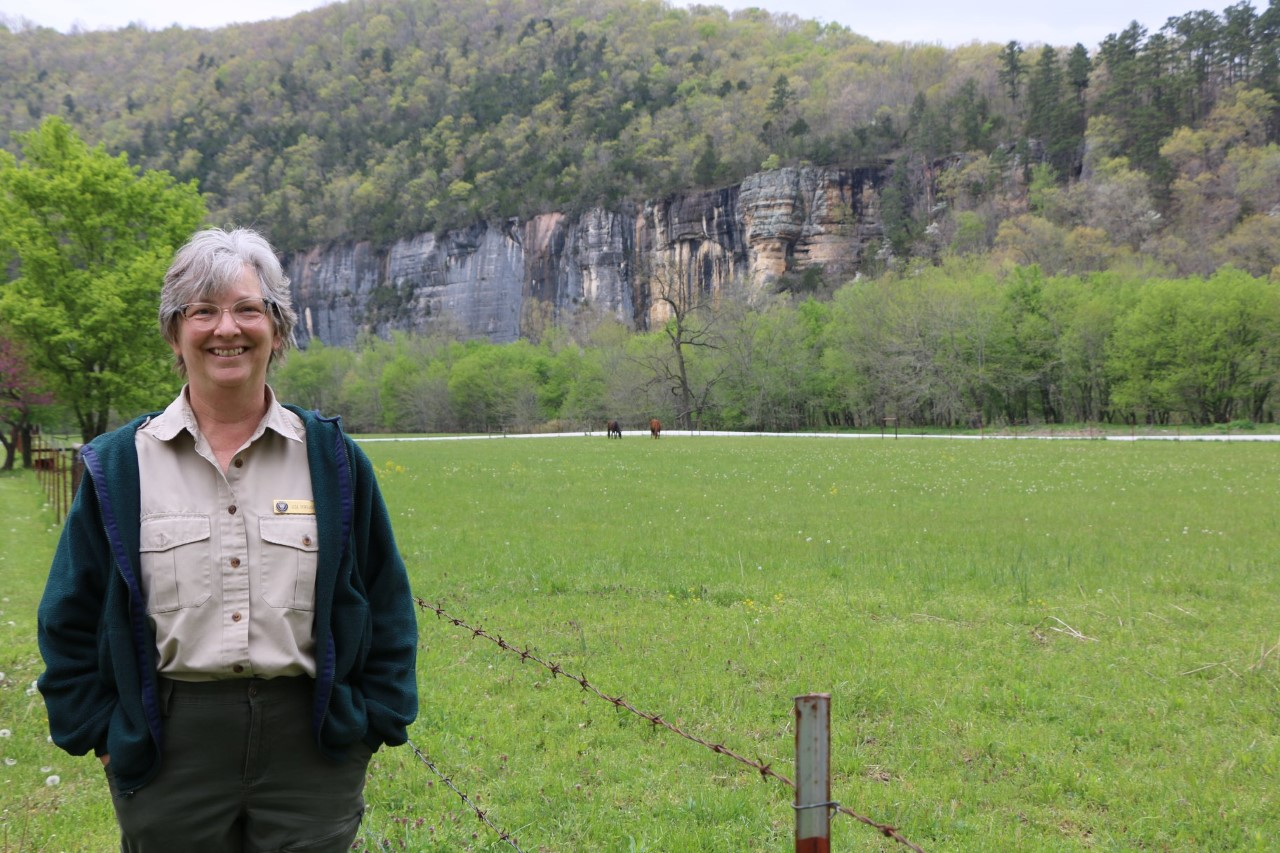 A woman wearing VIP uniform and a cardigan stands in front of a bluff.