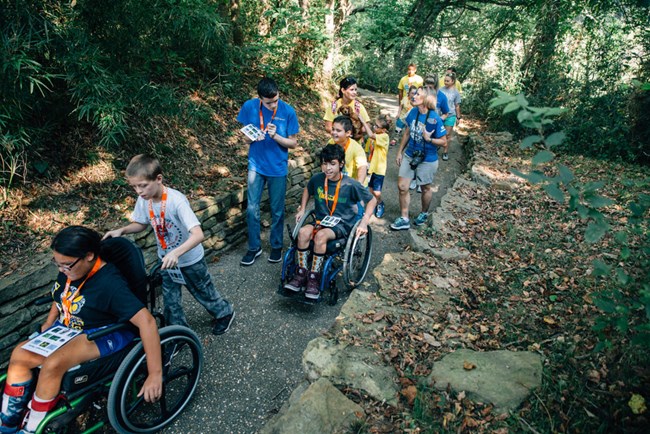 Students use wheelchairs on a paved path in the woods
