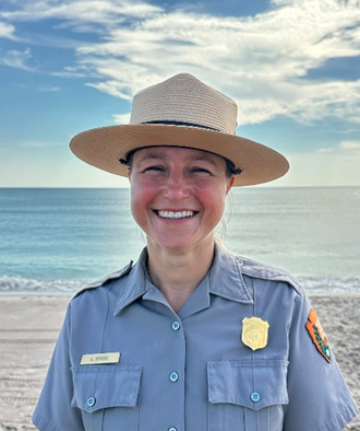 A Boyers Woman in a ranger hat smiling in front of a beach.