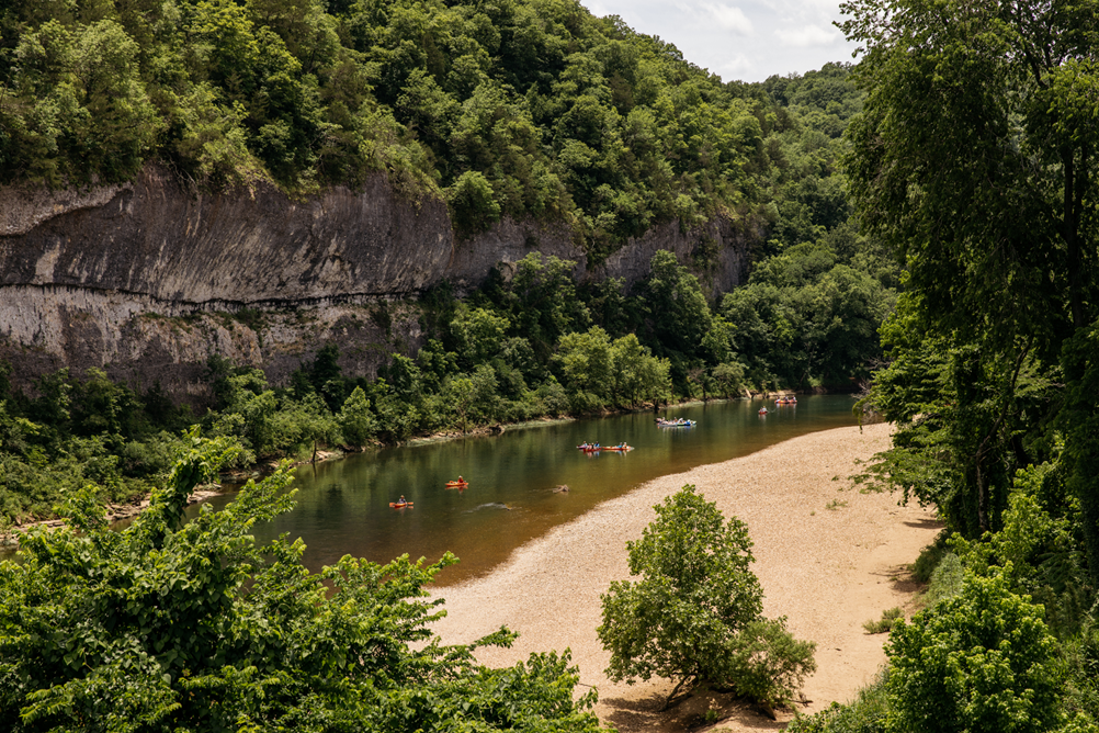 A section of Buffalo National River. A stretch of river, with several colorful kayaks on it, can be seen flowing at the base of a tan and gray streaked bluff. Tree covered hillsides can be seen flanking the far side of the river. A sandy gravel bar stretches the entire length of the river on