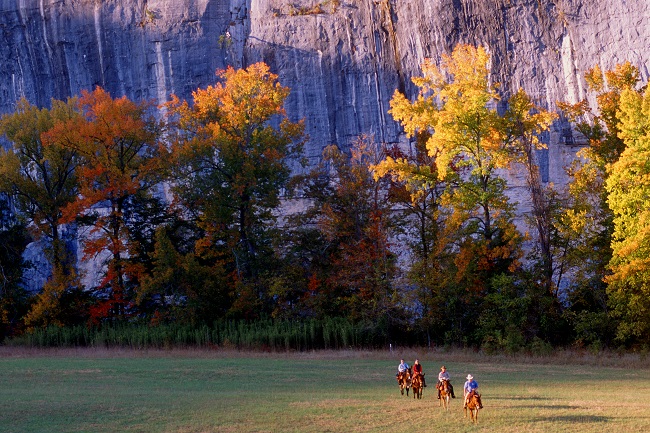 color photo with horseback riders on green grass at center bottom and trees with orange and yellow leaves with gray cliffs in background