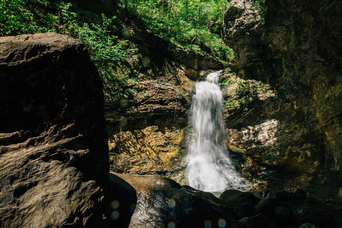 A color photograph of rugged steep tan rocks with a waterfall flowing over them and green trees in the background.