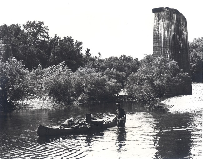 GilbertTrainBridgePier1967 - Neil Compton black and white photo of man walking in water behind canoe full of trash with shrubs and concrete tower remains of bridge support in background