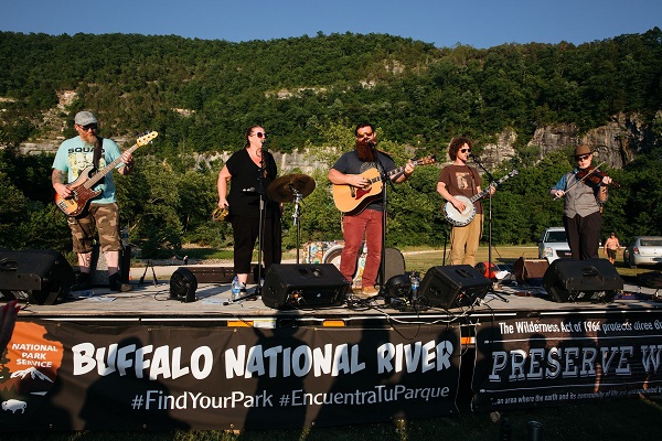 Five people on a stage playing instruments with tree covered mountain in background