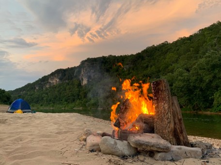An orange flamed fire within a ring of rocks on a sandy gravel bar can be seen in the forefront. A blue tent on the sand and green trees on a rocky bluff line can be seen in the background. Wispy white clouds are above the bluff line.