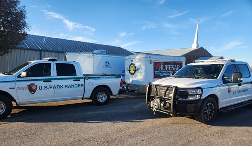Two U.S. Park Ranger pickup trucks are parked on pavement. Emergency service trailers can be seen in the background.