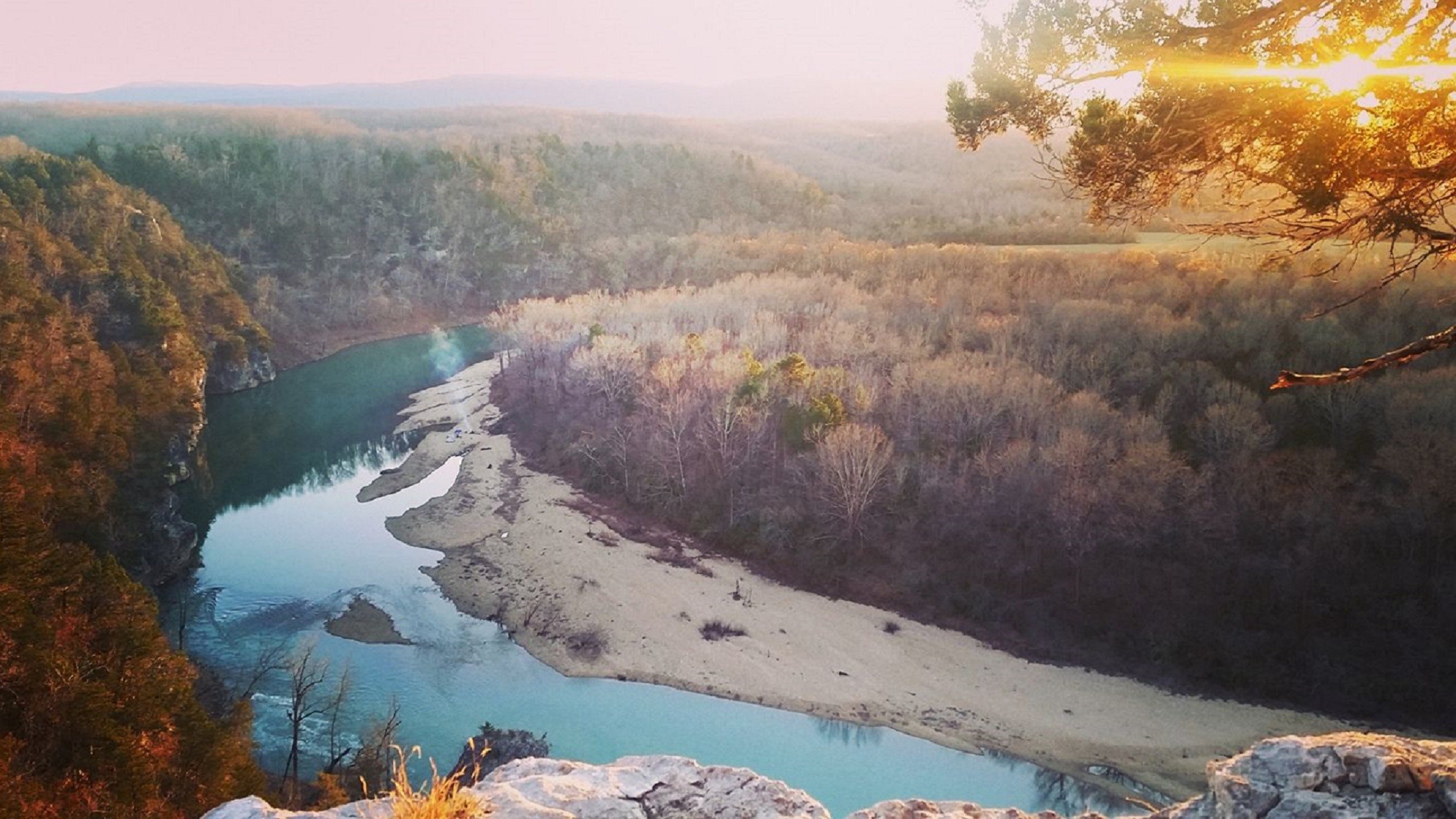 A scenic landscape showing a turquoise Buffalo National River winding through the forested hills of the Ozarks with the sun low on the horizon.