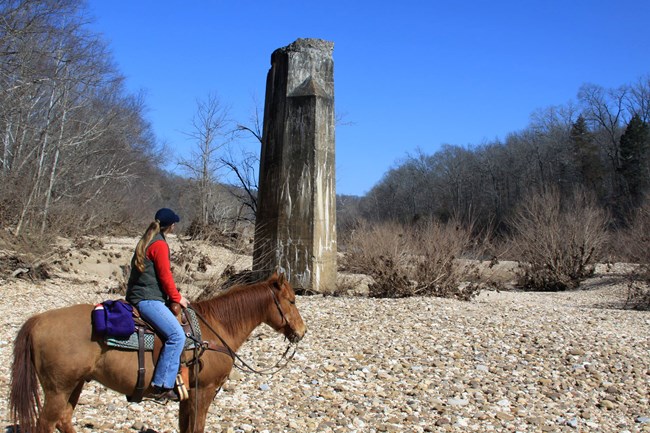 A horse and rider face the Buffalo River and a historic railroad bridge pier.