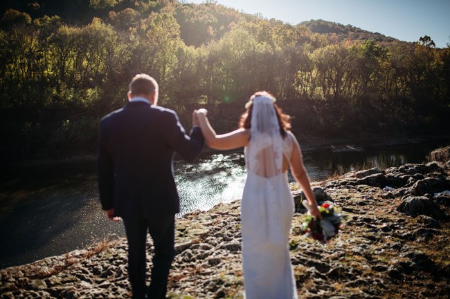 A bride and groom walk away from us while holding hands and looking over the river.